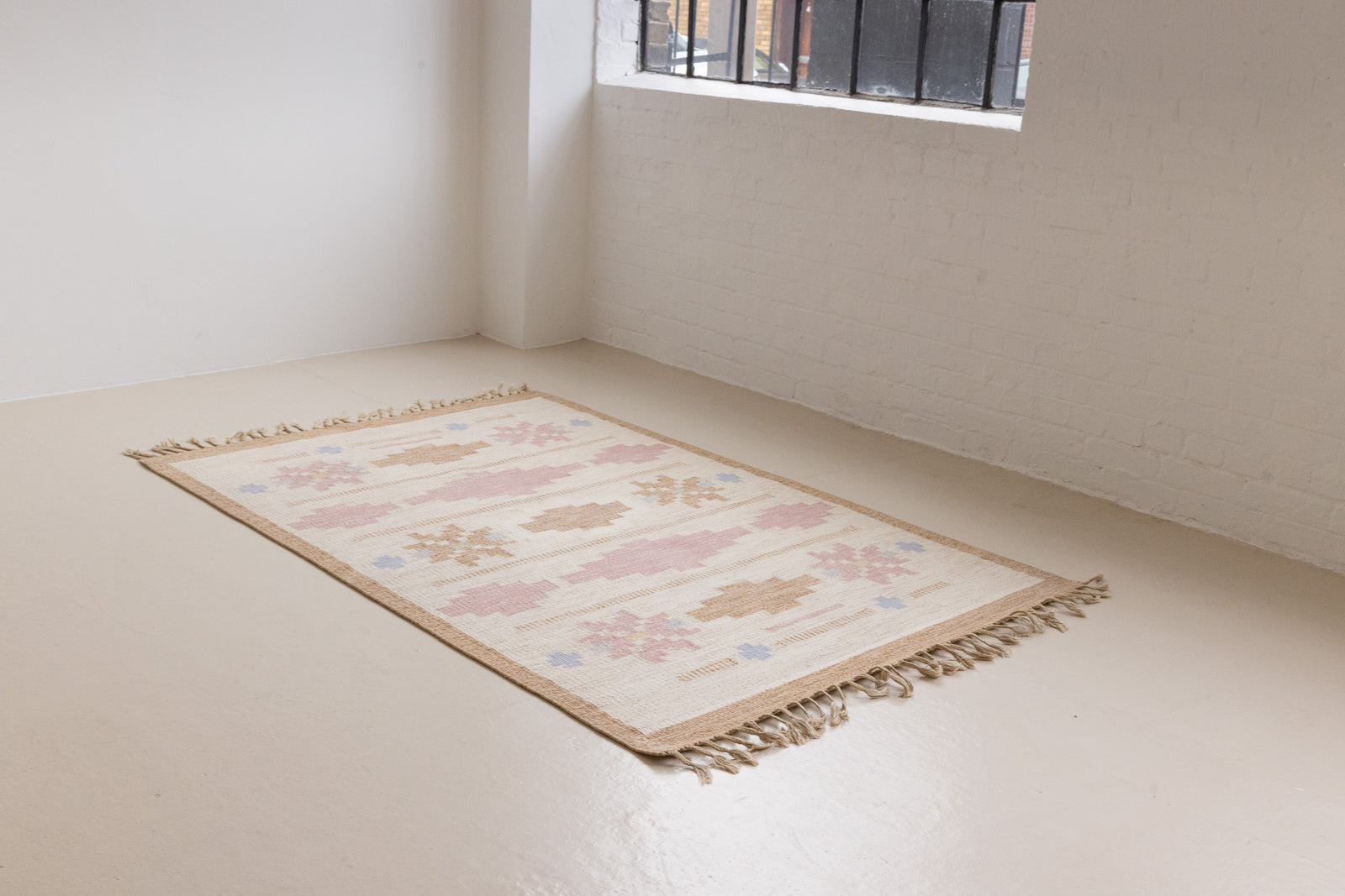 Trina is an original Swedish röllakan rug handwoven by the mid-century textile designer Anna Johanna Angström. This vintage rug features geometric designs in pink, blue, and brown on a beige ground.