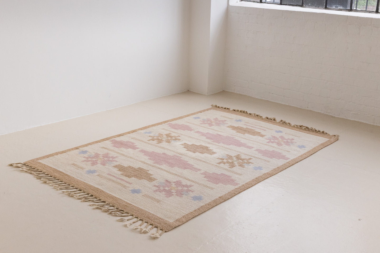 Trina is an original Swedish röllakan rug handwoven by the mid-century textile designer Anna Johanna Angström. This vintage rug features geometric designs in pink, blue, and brown on a beige ground.
