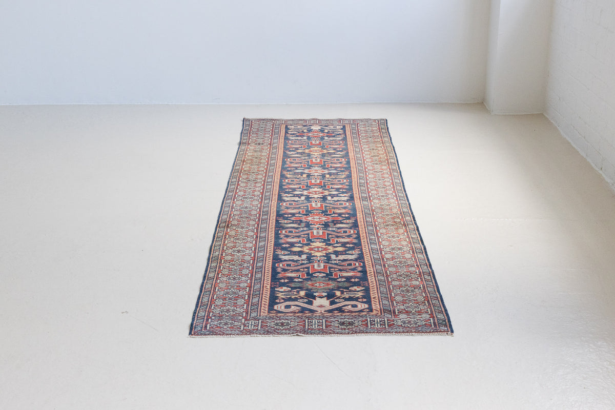 Beau is an antique Persian Caucasian runner in red and cream on a deep indigo ground, framed by a wide geometric border. Handwoven Caucasian carpets are prized for their bold geometric patterns and rich, saturated and jewel tones. 