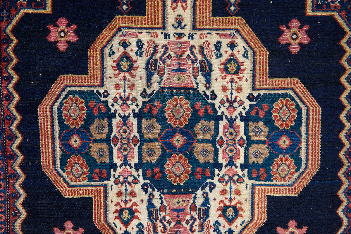 Close up of an antique handwoven Persian Senneh rug, with an intricate ivory medallion in pink and blue set on a deep indigo field. Persian Senneh carpets are known for their fine, intricate patterns and distinctive weaving technique.