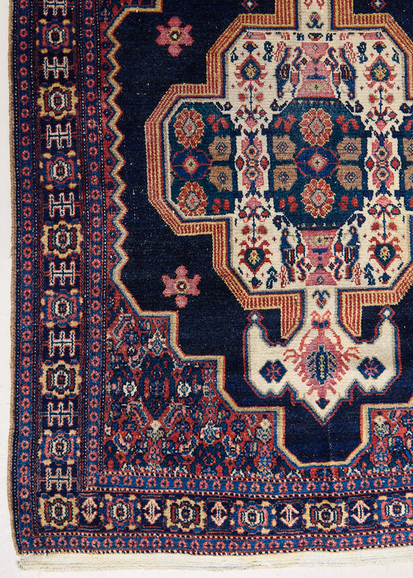 Belle is a small antique handwoven Persian Senneh rug, with an intricate ivory medallion in pink and blue set on a deep indigo field. Persian Senneh carpets are known for their fine, intricate patterns and distinctive weaving technique.