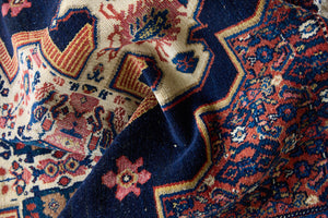 Close up of a small antique handwoven Persian Senneh rug, with an intricate ivory medallion in pink and blue set on a deep indigo field. Persian Senneh carpets are known for their fine, intricate patterns and distinctive weaving technique.