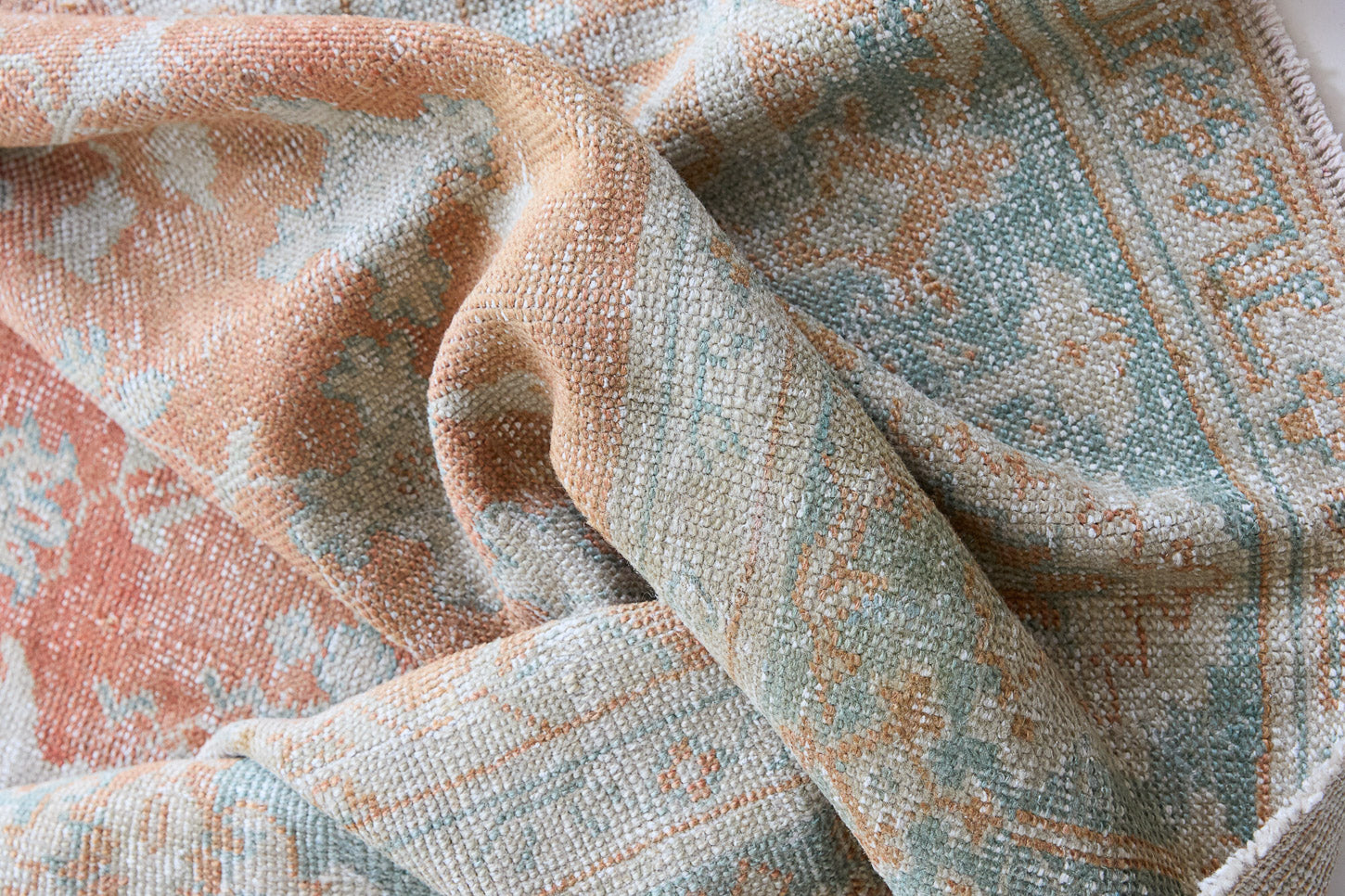 Close-up of textured fabric with a pattern in soft colors. An antique Turkish Oushak rug. Perdita features a large-scale, open medallion pattern, flowing botanical motifs and a generous field space set against a blue and terracotta ground.
