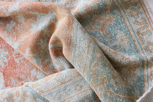 Close-up of textured fabric with a pattern in soft colors. An antique Turkish Oushak rug. Perdita features a large-scale, open medallion pattern, flowing botanical motifs and a generous field space set against a blue and terracotta ground.