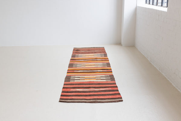 A South Anatolian nomadic kilim runner. Hand-woven on village and nomadic looms in vivid, naturally dyed colours, a tradition stretching back thousands of years. Vespa features a bold stripe pattern in earthy hues of rust, orange, taupe and black.