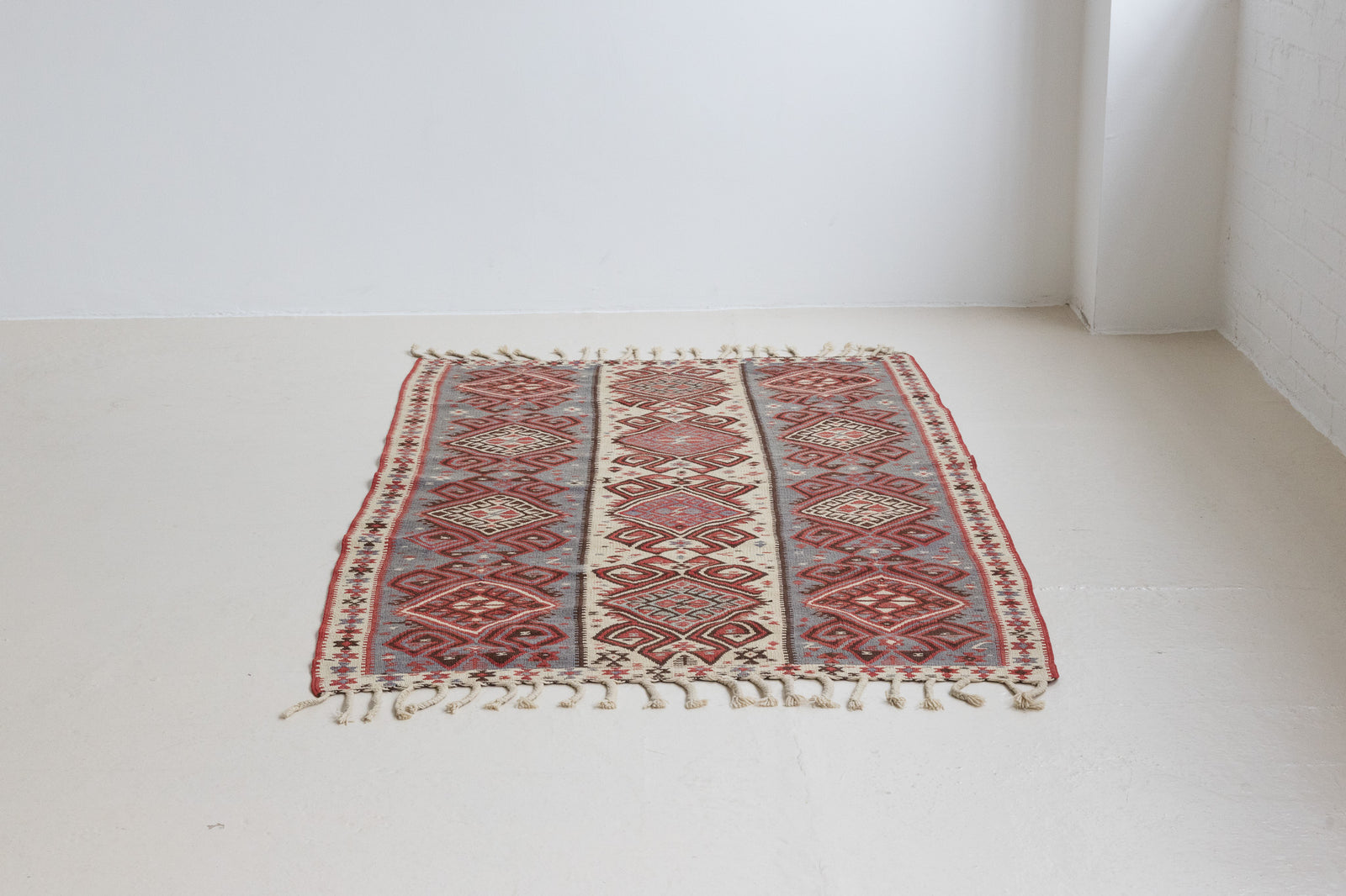 Zerrin is a bold, geometric Anatolian Hakkari kilim. Hand-woven on village and nomadic looms in vivid, naturally dyed colours, a tradition stretching back thousands of years. Kilims were woven for the practicalities of daily life and used as floor and wall coverings, storage bags, and prayer mats. Zerrin features a taupe and grey striped field scattered with the classic hand-on-hips motif, bordered by the comb motif both traditionally associated with marriage and fertility.