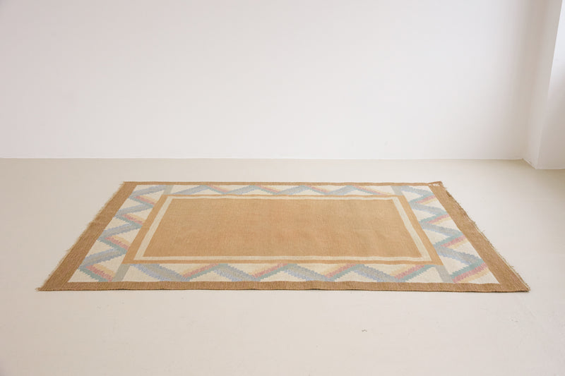A Swedish geometric-patterned rug with multicolor border on a beige background. This röllakan rug was handwoven by the mid-century textile designer Anna Johanna Angström.