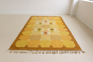 A vintage flatweave rug with geometric and floral patterns in yellow, beige, and other colors, with a brown border and white tassels. Barbara is a röllakan rug handwoven by designer Ingegerd Silow.
