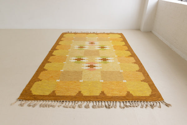 A vintage flatweave rug with geometric and floral patterns in yellow, beige, and other colors, with a brown border and white tassels. Barbara is a röllakan rug handwoven by designer Ingegerd Silow.