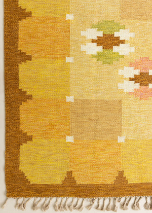 A vintage flatweave rug with geometric and floral patterns in yellow, beige, and other colors, with a brown border and white tassels. Barbara is a röllakan rug handwoven by designer Ingegerd Silow.