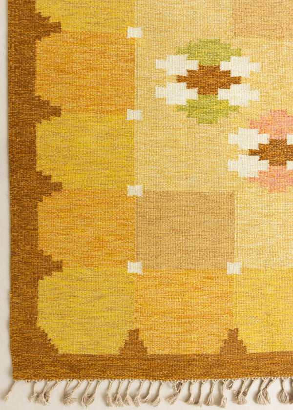 A vintage flatweave rug with geometric and floral patterns in yellow, beige, and other colors, with a brown border and white tassels. Barbara is a röllakan rug handwoven by designer Ingegerd Silow.