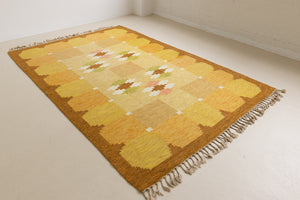 A vintage flatweave rug with geometric and floral patterns in yellow, beige, and other colors, with a brown border and white tassels. Barbara is a röllakan rug handwoven by designer Ingegerd Silow.