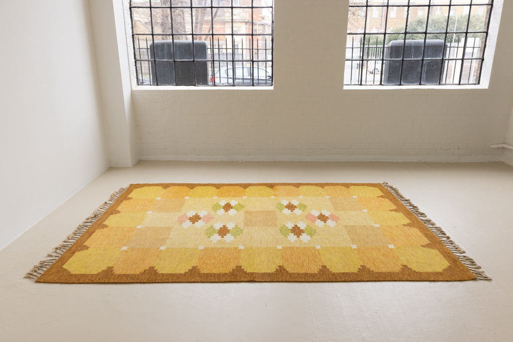 A vintage flatweave rug with geometric and floral patterns in yellow, beige, and other colors, with a brown border and white tassels. Barbara is a röllakan rug handwoven by designer Ingegerd Silow.