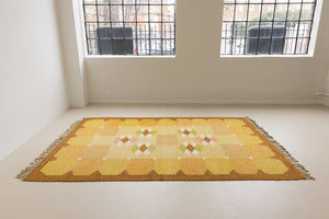A vintage flatweave rug with geometric and floral patterns in yellow, beige, and other colors, with a brown border and white tassels. Barbara is a röllakan rug handwoven by designer Ingegerd Silow.