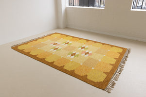 A vintage flatweave rug with geometric and floral patterns in yellow, beige, and other colors, with a brown border and white tassels. Barbara is a röllakan rug handwoven by designer Ingegerd Silow.