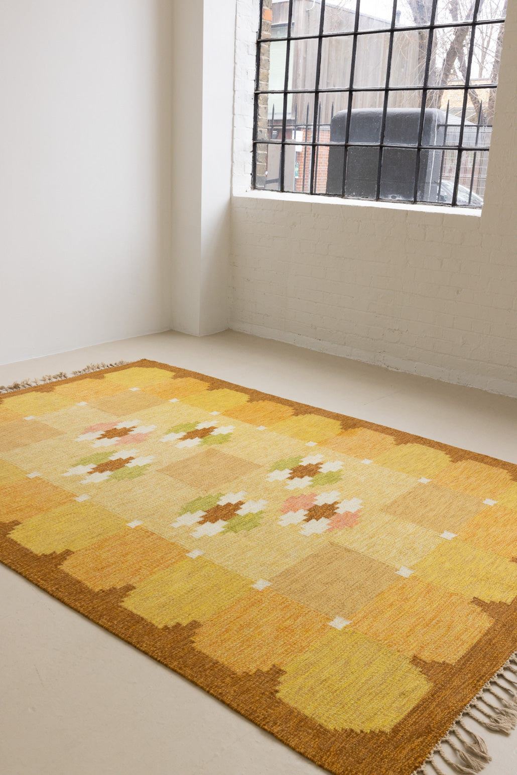 A vintage flatweave rug with geometric and floral patterns in yellow, beige, and other colors, with a brown border and white tassels. Barbara is a röllakan rug handwoven by designer Ingegerd Silow.