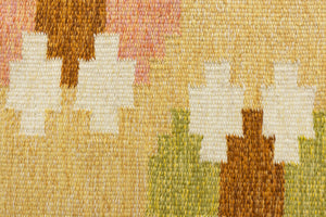 A vintage flatweave rug with geometric and floral patterns in yellow, beige, and other colors, with a brown border and white tassels. Barbara is a röllakan rug handwoven by designer Ingegerd Silow.