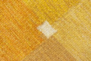 A vintage flatweave rug with geometric and floral patterns in yellow, beige, and other colors, with a brown border and white tassels. Barbara is a röllakan rug handwoven by designer Ingegerd Silow.
