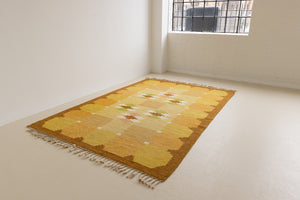 A vintage flatweave rug with geometric and floral patterns in yellow, beige, and other colors, with a brown border and white tassels. Barbara is a röllakan rug handwoven by designer Ingegerd Silow.